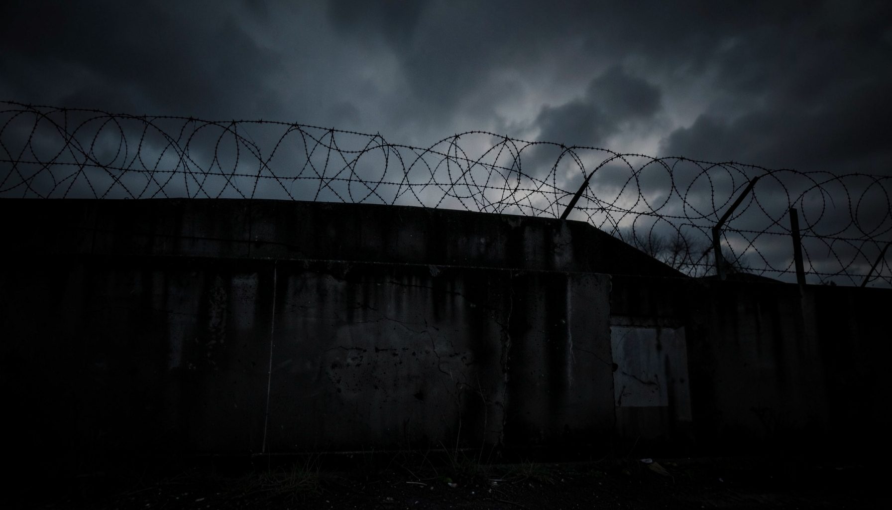 Dark atmospheric photograph of a prison wall with barbed wire against a moody sky, deep blacks and muted greys