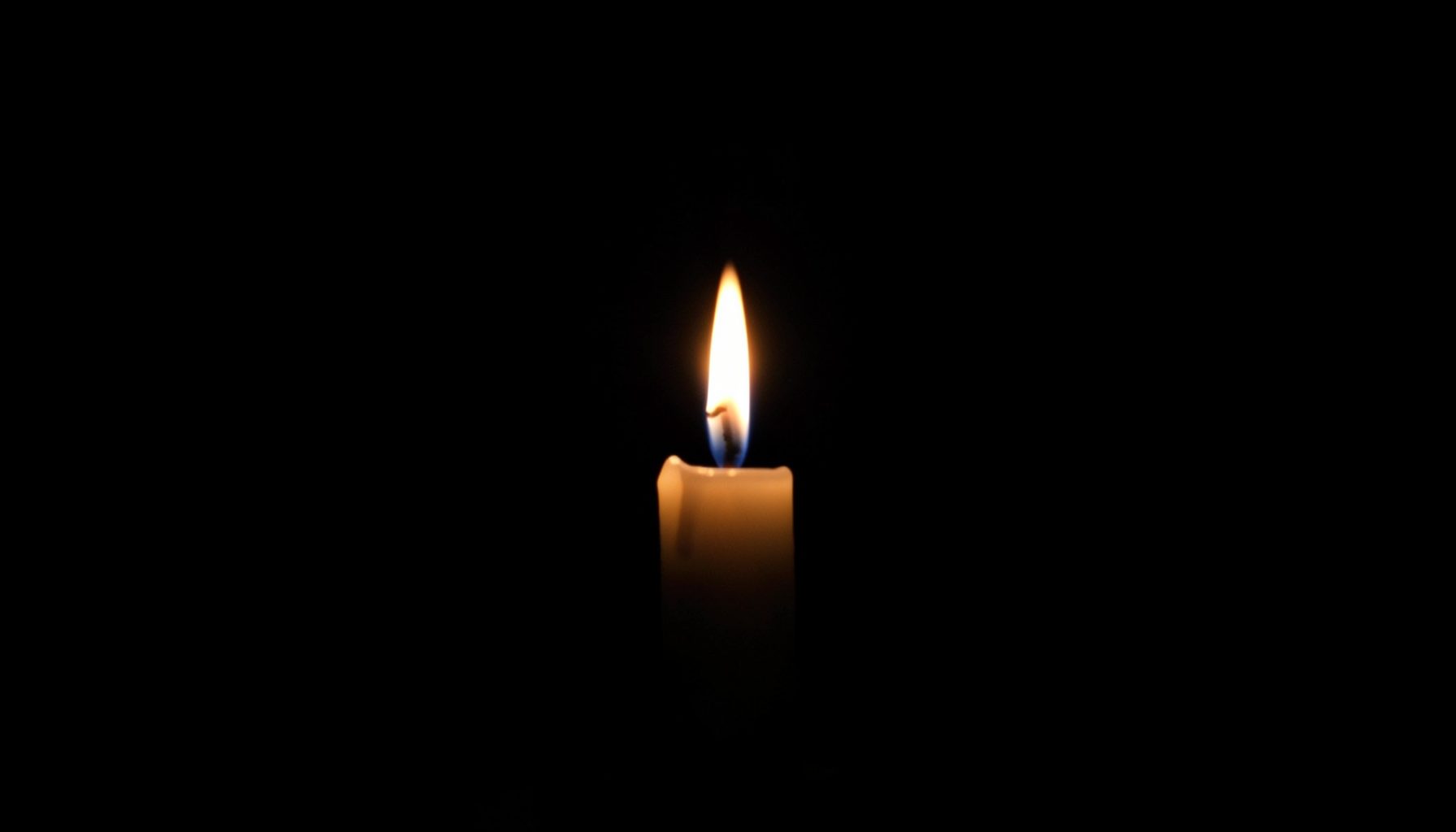 Dark atmospheric scene with a single candle flame glowing against a black background, symbolising remembrance and solemnity