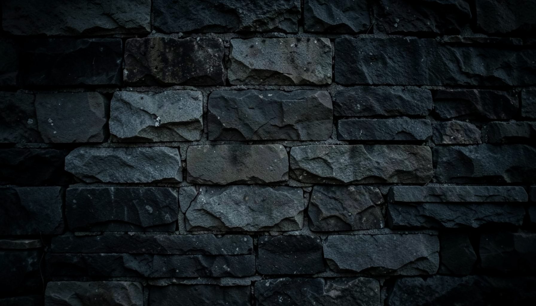 Dark, moody photograph of a stone wall with muted grey and charcoal tones, evoking a sombre and reflective atmosphere
